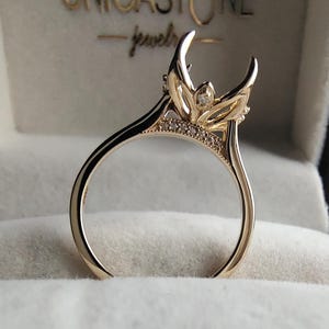 Op de afbeelding: Een gouden ringzetting met een decoratieve kroonvormige kop met gebogen pootjes en kleine pavé-gezette steentjes langs de brug. De ring wordt getoond in een doosje met de tekst UNICASTONE jewels in goud gedrukt op het binnendeksel.
