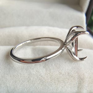 May include: A polished silver-toned metal ring setting featuring a smooth, thin band and a decorative, multi-pronged head designed to hold a gemstone. The setting is empty, showcasing the intricate metalwork of the prongs and the band's profile.