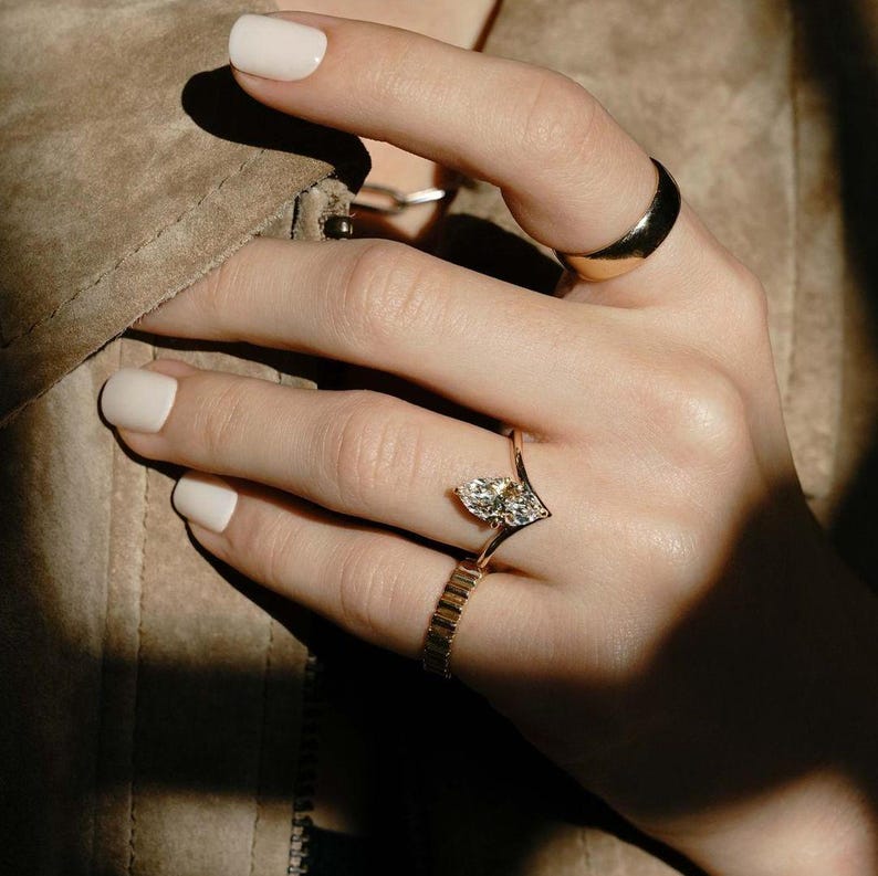 May include: A pear-shaped diamond engagement ring set in a gold band with a V-shaped design. The ring is worn on a finger with a gold band on the ring finger.