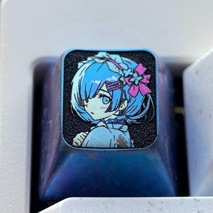 May include: A blue and black keyboard keycap with a colorful anime character illustration. The keycap features a female character with blue hair and a pink flower in her hair. The character is wearing a blue and white dress. The keycap is on a white keyboard.