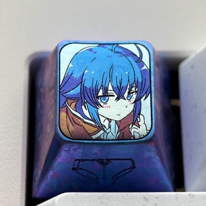 May include: A blue and purple keyboard keycap with a printed image of an anime character with blue hair. The character is wearing a brown and white outfit and has a pink blush on their cheeks. The keycap is labeled with the word "Tab" in white text.