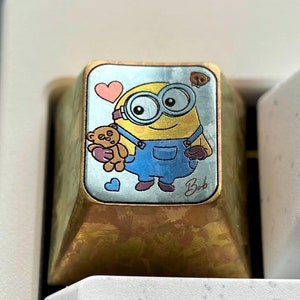 May include: A gold-colored keycap with a blue and yellow cartoon character holding a teddy bear. The character has a blue jumpsuit and goggles. The keycap is labeled "Bob".