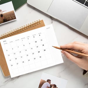 Pode incluir: Um calend&aacute;rio de mesa com espiral para julho de 2027 com capa castanha, apresentando uma grelha branca com texto preto para as datas e dias da semana. O calend&aacute;rio est&aacute; posicionado numa superf&iacute;cie branca ao lado de um computador port&aacute;til.