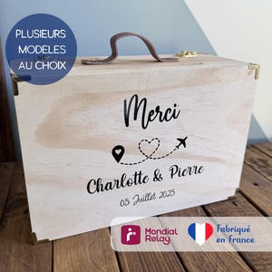 Peut inclure: Une boîte rectangulaire en bois clair avec une poignée marron et des coins métalliques. Le devant présente le texte noir Merci, Charlotte & Pierre, 05 Juillet 2025, ainsi qu'un motif de point de localisation, un cœur et un avion.