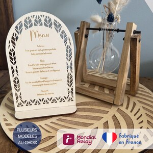 Peut inclure: Un menu en bois en forme d'arche avec des motifs de feuilles découpés au laser sur le bord, affichant le mot Menu et du texte. Il repose sur un socle circulaire en bois avec un motif de feuille assorti, accompagné d'un vase en verre.