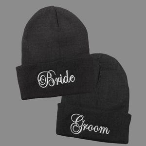 May include: Two black knit beanies with white embroidered text. One beanie says "Bride" and the other says "Groom".