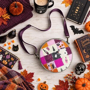 May include: A round crossbody bag featuring a patchwork design with various Halloween-themed illustrations including ghosts, witches, and potions in shades of pink, purple, orange, and black, equipped with an adjustable dark purple shoulder strap.