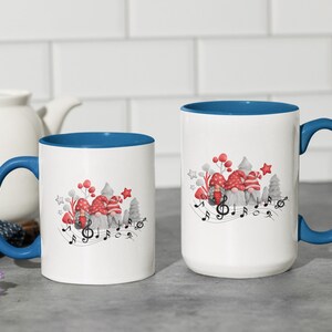 May include: Two white ceramic mugs with blue rims. The mugs feature a whimsical design of gnomes wearing red hats and singing with musical notes. The design is in watercolour style.