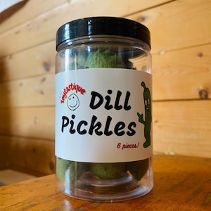 May include: A clear plastic jar filled with green dill pickle spears. The jar has a black lid and a white label that says "Toytastique Dill Pickles 6 pieces!" with a cartoon pickle on it.