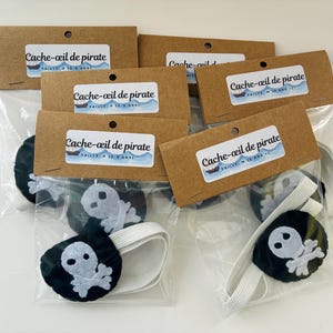 May include: Several pirate eye patches are displayed in clear plastic bags, each with a brown cardboard tag. The eye patches are black with a white skull and crossbones design. The tags read "Cache-œil de pirate".