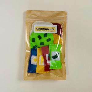 May include: A clear plastic bag containing a set of felt food items for pretend play. The set includes a green felt burger bun with green felt lettuce and a red felt burger patty. There are also five felt condiment packets in yellow, blue, red, and green.