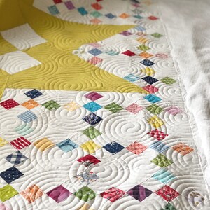 May include: A white quilt with a yellow star design and a patchwork of colorful squares. The quilt is quilted with a swirling pattern.