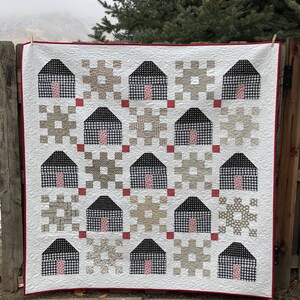 May include: A white quilt with a red border featuring a repeating pattern of black and white houses with pink doors. The houses are surrounded by various geometric patterns in shades of brown and beige.
