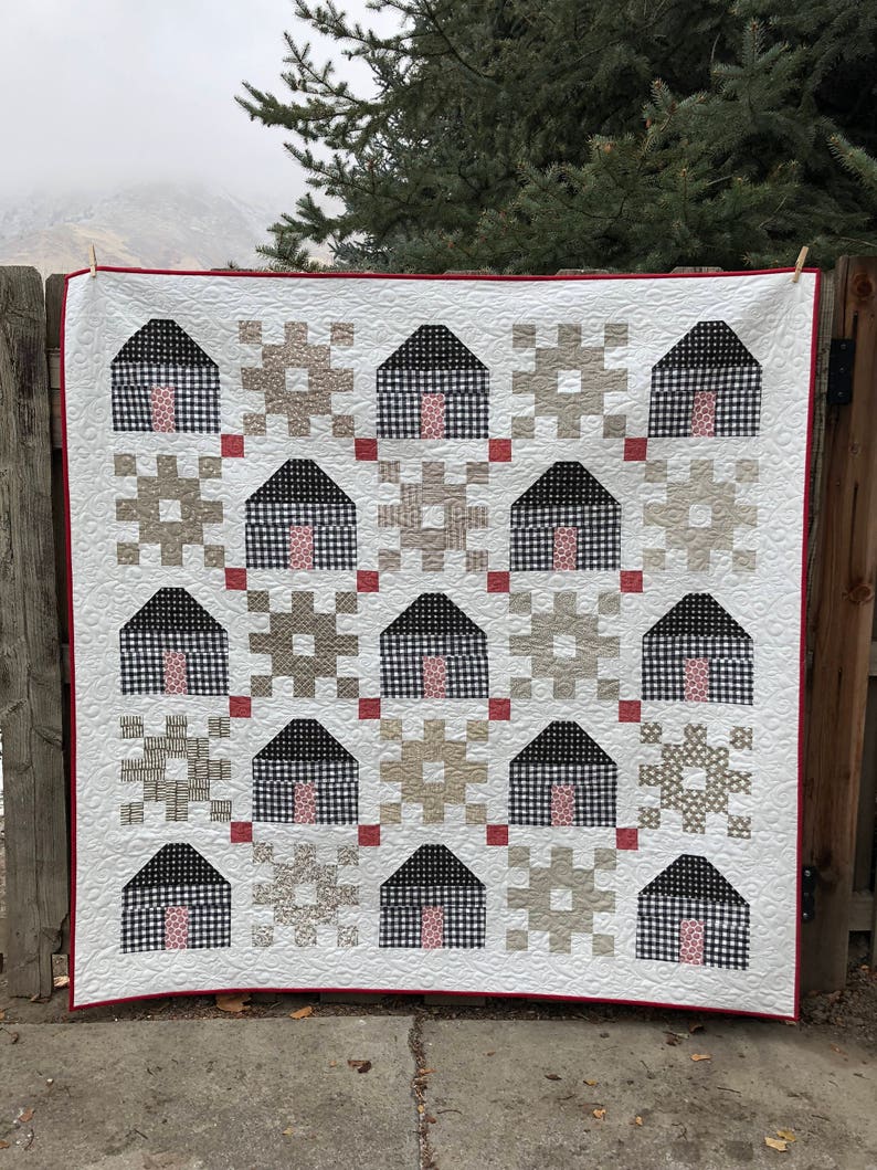May include: A white quilt with a red border featuring a repeating pattern of black and white houses with pink doors. The houses are surrounded by various geometric patterns in shades of brown and beige.
