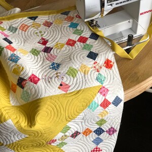 May include: A white quilt with a yellow border and a colorful patchwork design is being sewn on a Bernina sewing machine. The quilt has a swirling pattern of quilting stitches.