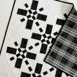 May include: Black and white quilt with a geometric pattern. The quilt is made of squares and rectangles, and the pattern is repeated throughout the quilt. The quilt is folded in half, and the back of the quilt is a black and white plaid.