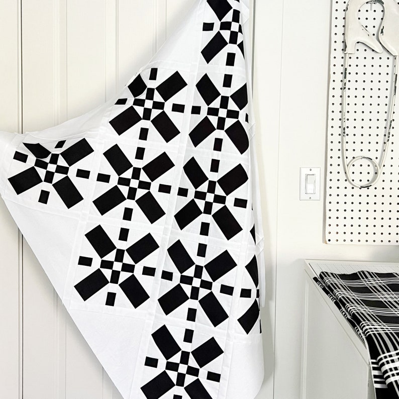 May include: A white fabric quilt with a black and white geometric pattern. The quilt is folded in half and hanging on a white door.