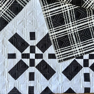 May include: Black and white quilt with a geometric pattern. The quilt is folded in half, showing the back side, which is a black and white plaid pattern.