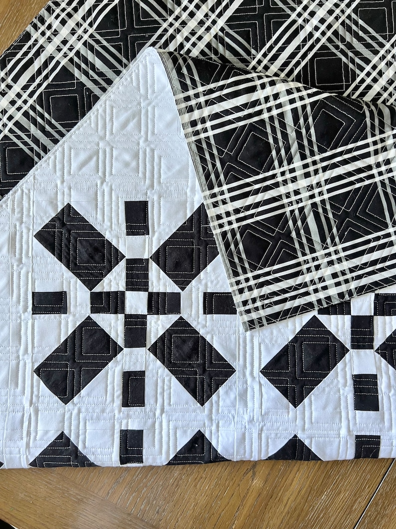 May include: Black and white quilt with a geometric pattern. The quilt is folded in half, showing the back side, which is a black and white plaid pattern.