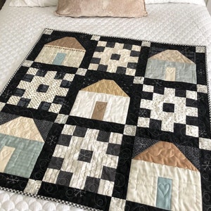 May include: A black and white quilt with a patchwork design featuring houses with brown roofs and blue doors. The quilt is made up of squares with different patterns, including checkered, polka dot, and floral.