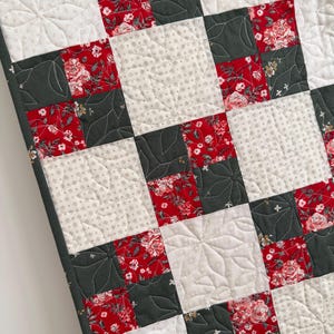 Może przedstawiać: Narzuta typu patchwork z powtarzającym się wzorem siatki z kwadratowych blok&oacute;w materiału. Projekt składa się z naprzemiennych kwadrat&oacute;w w kolorze ciemnego antracytu, czerwonego wzoru w kwiaty oraz kremowego materiału w gwiazdki.