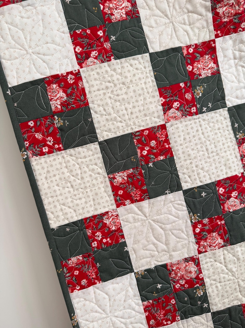 Może przedstawiać: Narzuta typu patchwork z powtarzającym się wzorem siatki z kwadratowych blok&oacute;w materiału. Projekt składa się z naprzemiennych kwadrat&oacute;w w kolorze ciemnego antracytu, czerwonego wzoru w kwiaty oraz kremowego materiału w gwiazdki.