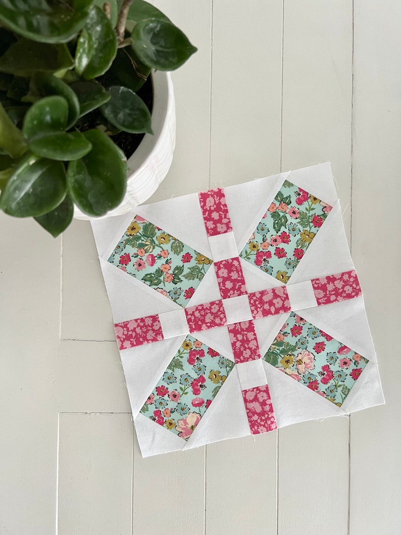 May include: A white quilt block with a pink and blue floral pattern. The block is made up of squares and triangles, and the pattern is repeated throughout the block.