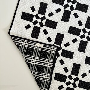 May include: Black and white quilt with a geometric pattern. The quilt is made of squares and rectangles, and the pattern is repeated throughout the quilt. The quilt is folded in half, and the back side of the quilt is a black and white plaid.