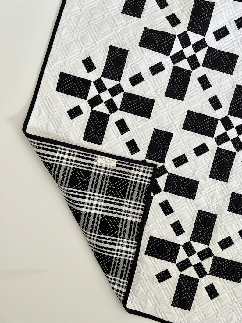 May include: Black and white quilt with a geometric pattern. The quilt is made of squares and rectangles, and the pattern is repeated throughout the quilt. The quilt is folded in half, and the back side of the quilt is a black and white plaid.