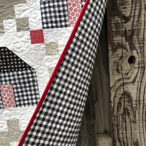 May include: A black and white gingham quilt with a red binding. The quilt is folded and leaning against a wooden fence.