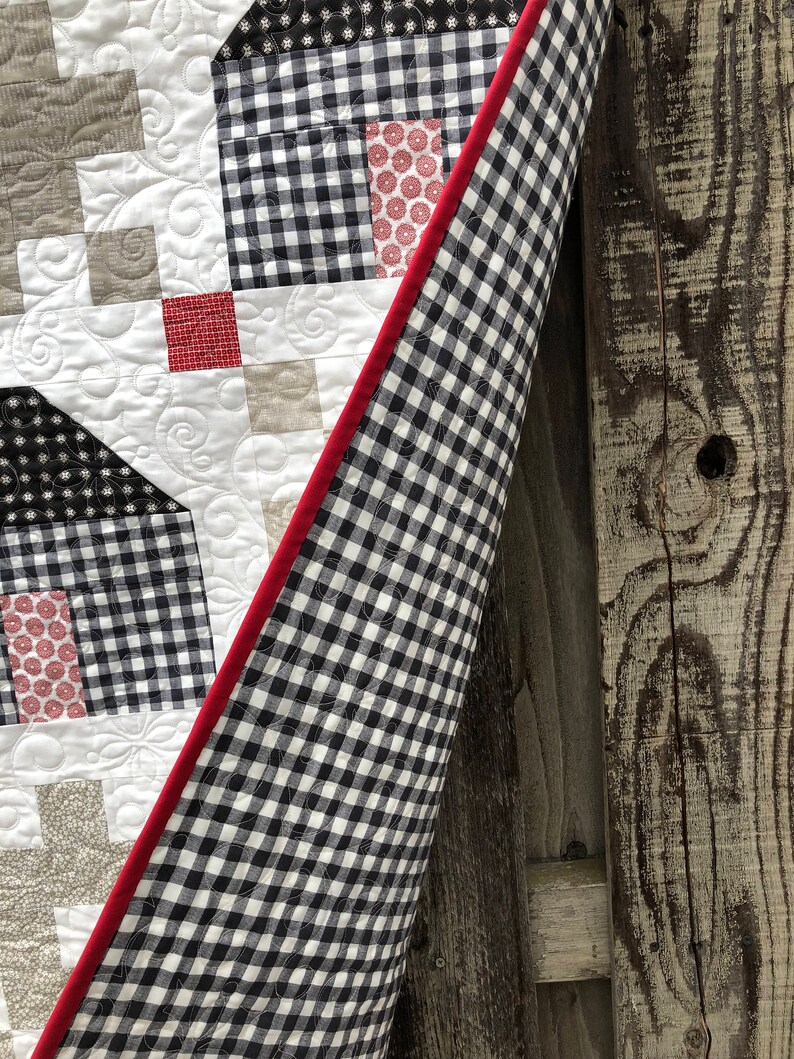 May include: A black and white gingham quilt with a red binding. The quilt is folded and leaning against a wooden fence.