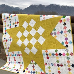May include: A yellow and white quilt with a star design in the center. The quilt is made up of small squares of fabric in various colors and patterns. The quilt is displayed against a backdrop of mountains.