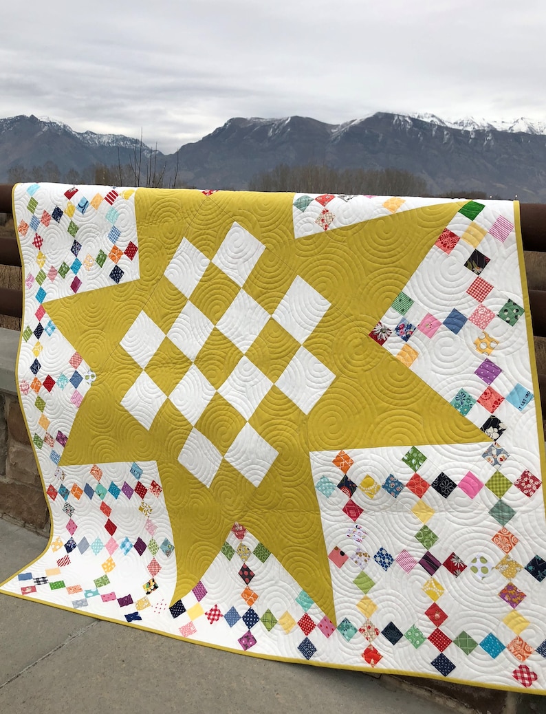 May include: A yellow and white quilt with a star design in the center. The quilt is made up of small squares of fabric in various colors and patterns. The quilt is displayed against a backdrop of mountains.