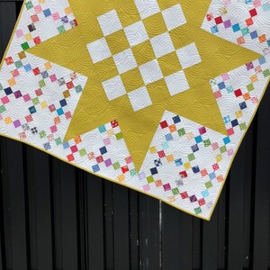 May include: A yellow and white quilt with a star design. The quilt is made of small squares of fabric in various colors and patterns. The quilt is hanging on a black fence.