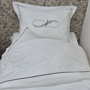 May include: White bedding with a black trim and a white pillow with a black infinity symbol embroidered on it.