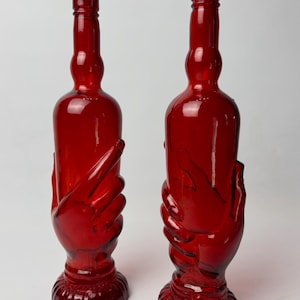 Peut inclure: Deux grandes bouteilles en verre rouge translucide, chacune présentant un motif moulé d'une main s'enroulant autour de la base et du bas du corps. Les bouteilles ont un col fin, une épaule arrondie et une base circulaire nervurée.