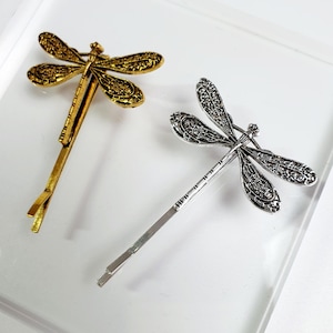 May include: Two dragonfly hair clips. One is gold-coloured, the other is silver-coloured. Both clips feature detailed wing designs and are positioned on a white surface. The dragonfly hair clips are decorative accessories.