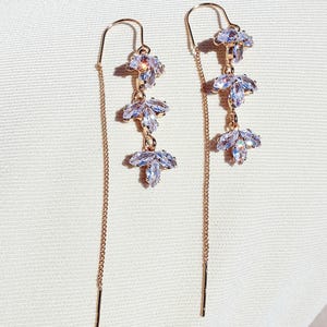 Pode incluir: Um par de brincos de fio em ouro rosa. Cada brinco apresenta três cristais claros e brilhantes em forma de folha. Os brincos têm uma corrente delicada e uma barra reta na extremidade. O fundo é um tecido claro esbranquiçado.