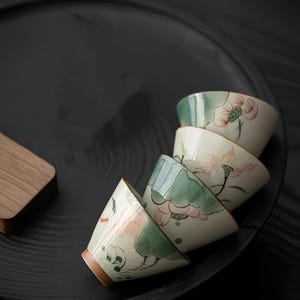 Puede incluir: Un juego de cuatro tazas de té de cerámica blanca con diseños florales verdes y rosas. Las tazas están apiladas una encima de la otra sobre una superficie negra.