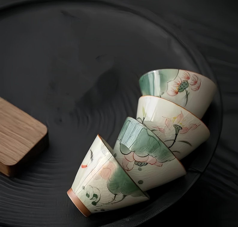 May include: A set of four white ceramic teacups with green and pink floral designs. The cups are stacked on top of each other on a black surface.