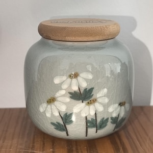 May include: A ceramic jar with a light blue glaze and a wooden lid. The jar is decorated with white flowers and green leaves.