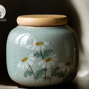 May include: A ceramic jar with a light blue glaze and a wooden lid. The jar is decorated with white flowers and green leaves.