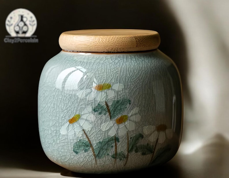 May include: A ceramic jar with a light blue glaze and a wooden lid. The jar is decorated with white flowers and green leaves.