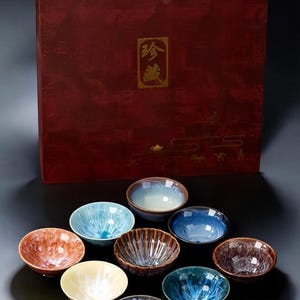 May include: A set of ten small, colorful ceramic teacups in a wooden box. The box is red with gold lettering that reads "香". The teacups are arranged in a circle on a gray surface.