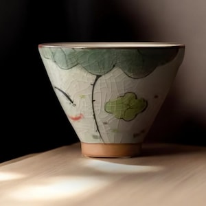 May include: A white ceramic teacup with a green and brown floral design. The cup has a wide, rounded base and a slightly flared rim.