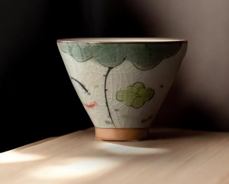 May include: A white ceramic teacup with a green and brown floral design. The cup has a wide, rounded base and a slightly flared rim.