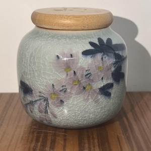 May include: A ceramic jar with a wooden lid, featuring a floral design with pink and blue flowers on a light blue background.