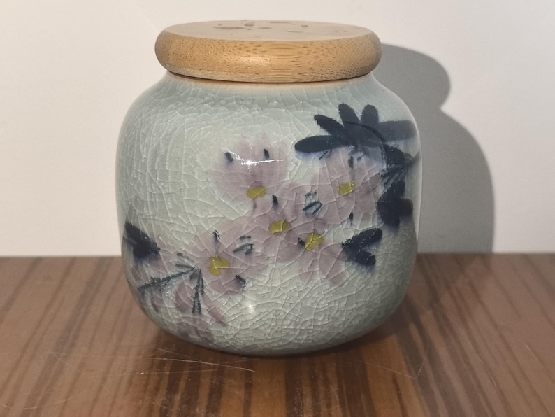 May include: A ceramic jar with a wooden lid, featuring a floral design with pink and blue flowers on a light blue background.