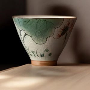 May include: A white ceramic teacup with a green and red floral design. The cup has a wide, rounded shape and a slightly flared lip.