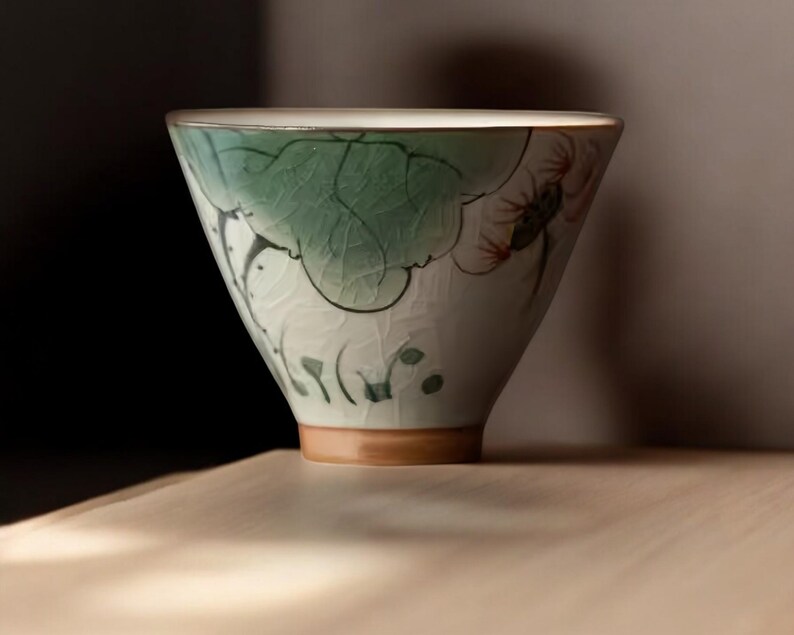 May include: A white ceramic teacup with a green and red floral design. The cup has a wide, rounded shape and a slightly flared lip.